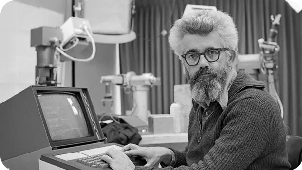 Foto de uma das primeiras pessoas a usar o termo Inteligência Artificial, John McCarthy, professor de matemática do Dartmouth College. Na imagem, temos uma foto de McCarthy, um homem branco, com barba grande, cabelos grisalhos e um óculos de grau com armação preta digitando em um antigo computador enquanto olha para a câmera.